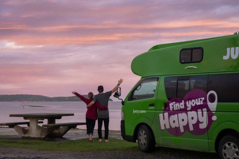 Two people watch the sunset from their jucy campervan by the ocean New Zealand